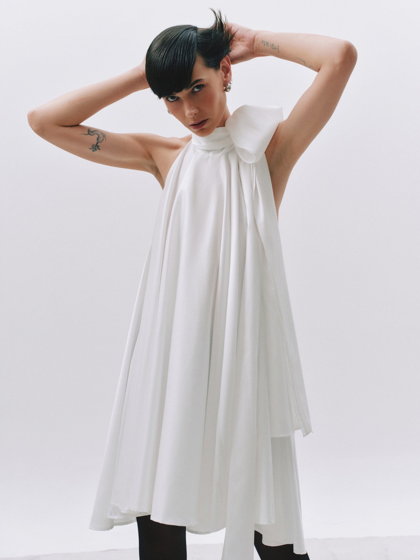 A model with short black hair poses in a sleeveless white dress featuring a large bow at the neckline, set against a plain light gray studio background.