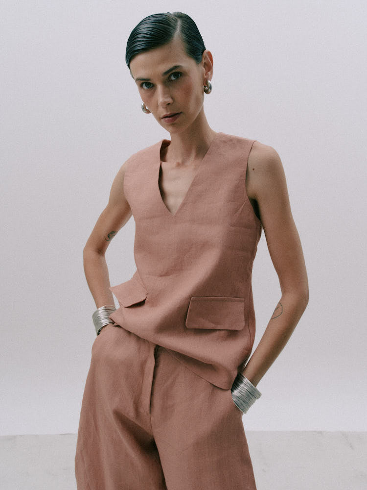 A model wearing a dusty rose linen two-piece set consisting of a sleeveless V-neck top and matching wide-leg trousers, styled with silver cuff bracelets against a plain light gray background.