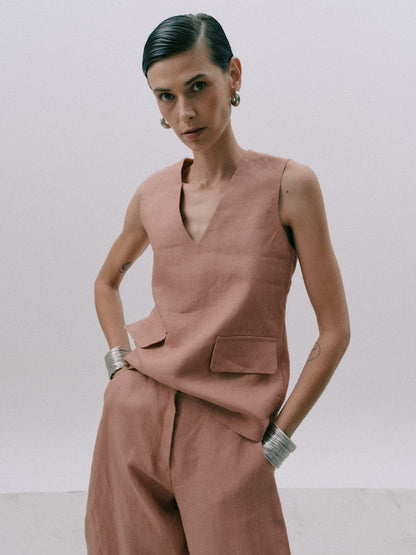 A model wearing a dusty rose linen two-piece set consisting of a sleeveless V-neck top and matching wide-leg trousers, styled with silver cuff bracelets against a plain light gray background.
