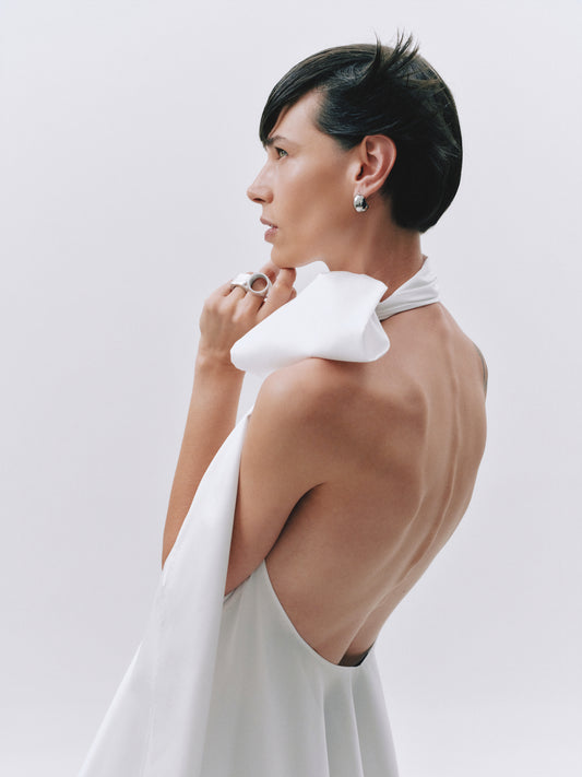 A model with short dark hair is shown from the back and side, wearing a white halter dress with a large draped collar and a low open back. She is wearing a silver ring and earring against a plain light gray studio background.