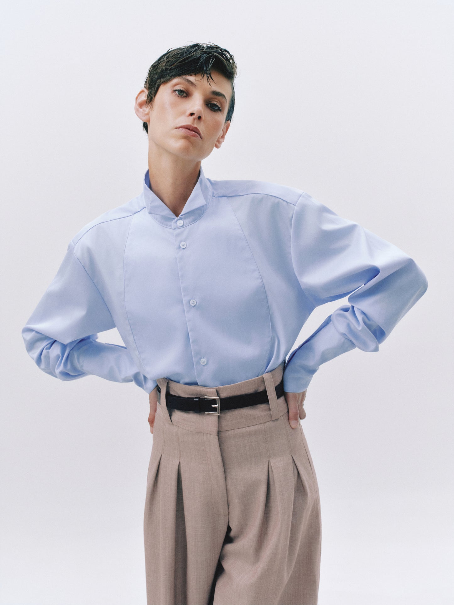 A model wearing a light blue long-sleeve button-up shirt with a unique pleated front and a stand collar, paired with beige pleated trousers and a black belt, against a light gray studio background.