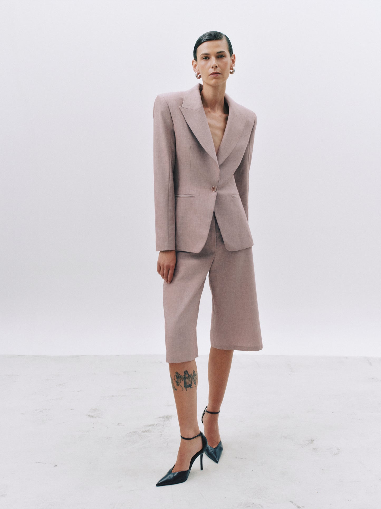A model stands in a studio wearing a tailored two-piece suit in a muted dusty pink or mauve tone, with matching cropped blazer and knee-length shorts. The outfit is styled with dark pointed-toe heels and hoop earrings against a clean light gray background.