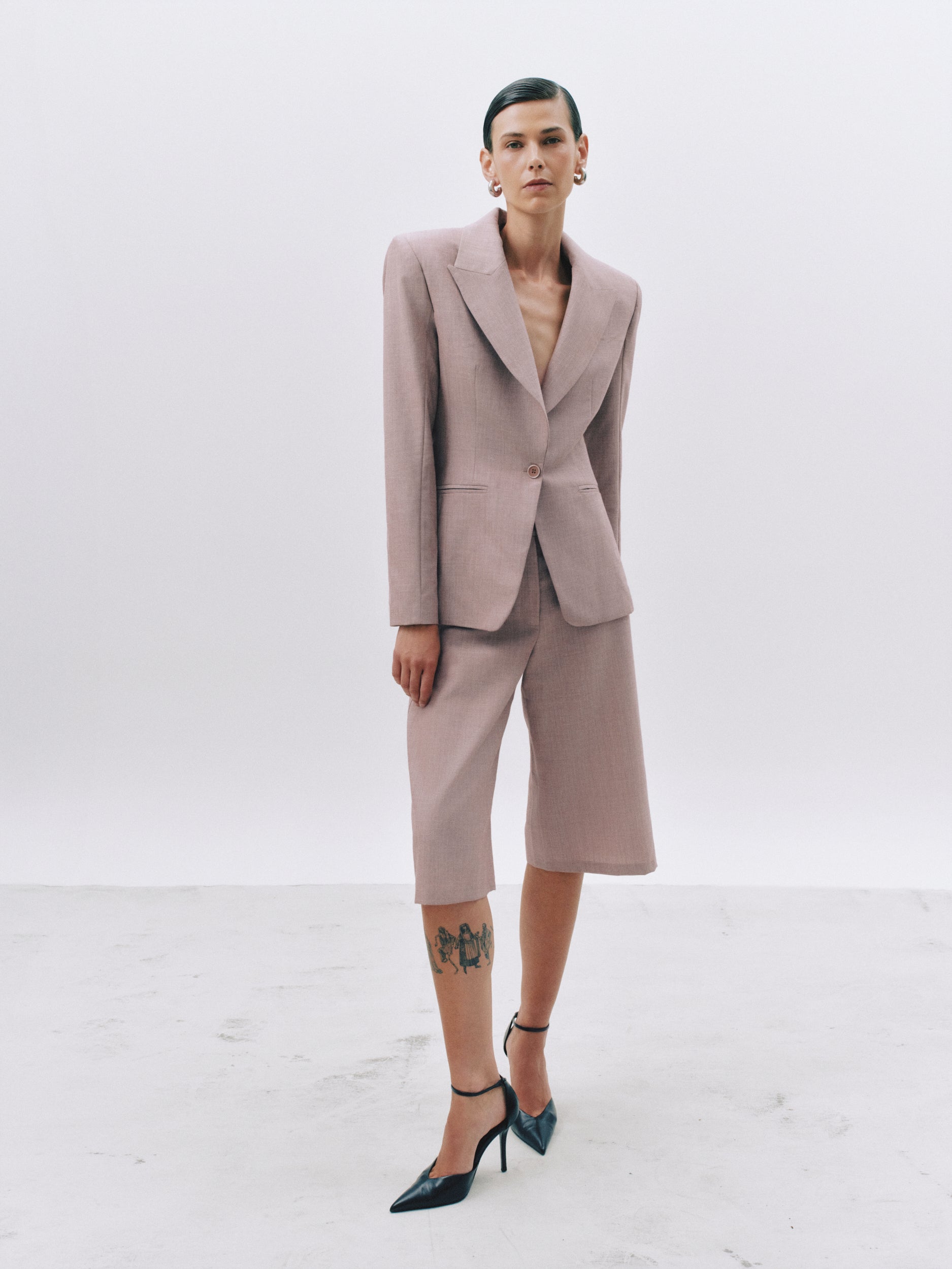 A model stands in a studio wearing a tailored two-piece suit in a muted dusty pink or mauve tone, with matching cropped blazer and knee-length shorts. The outfit is styled with dark pointed-toe heels and hoop earrings against a clean light gray background.