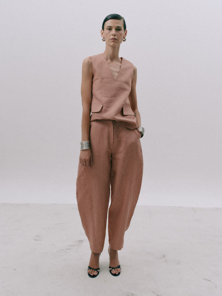 A model stands against a plain light gray studio background wearing a coordinated dusty rose outfit consisting of a sleeveless V-neck top with flap pockets and matching loose wide-leg pants, paired with black strappy sandals.
