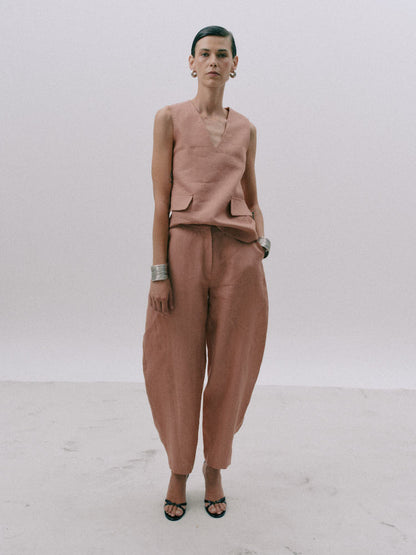 A model stands against a plain light gray studio background wearing a coordinated dusty rose outfit consisting of a sleeveless V-neck top with flap pockets and matching loose wide-leg pants, paired with black strappy sandals.